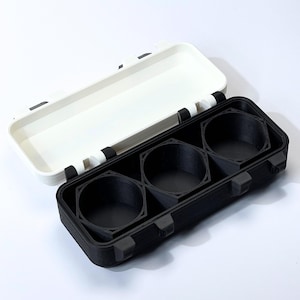 May include: A black and white rectangular case with a hinged lid. The case is open, revealing three circular compartments. The lid is white, and the base is black. The case appears to be made of plastic.