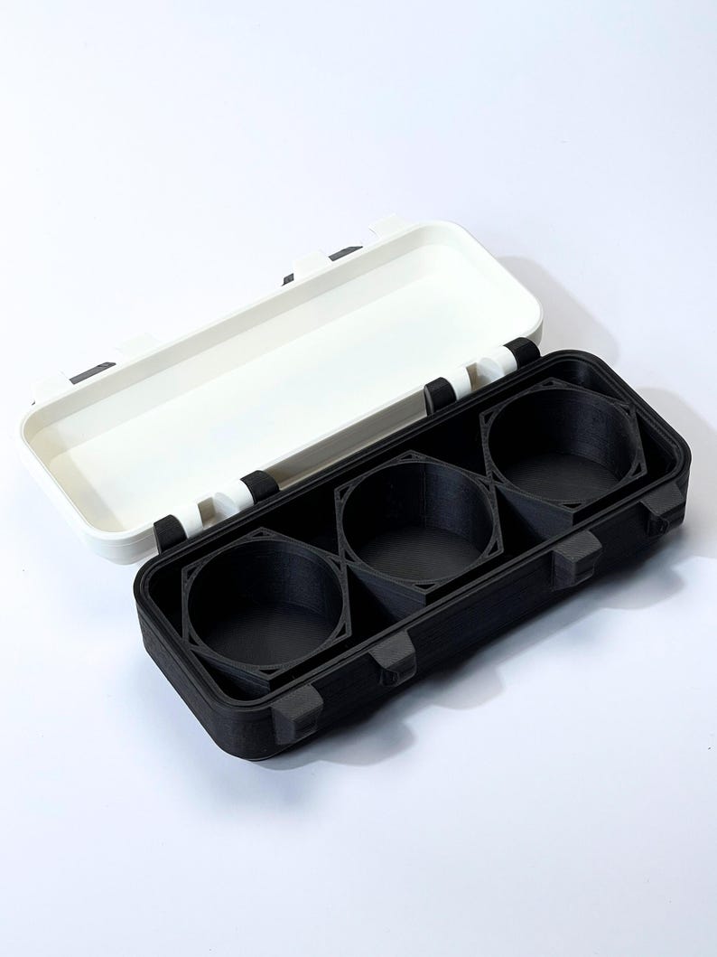 May include: A black and white rectangular case with a hinged lid. The case is open, revealing three circular compartments. The lid is white, and the base is black. The case appears to be made of plastic.