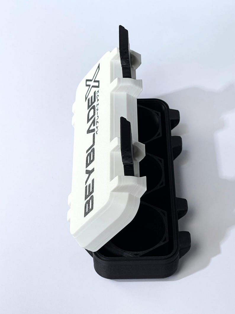 May include: A white and black plastic case with the text "BEYBLADEX" printed on the lid. The case is open, revealing black circular compartments. The case is designed to hold small items.