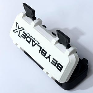 May include: A white and black plastic case with the text "BEYBLADE X" printed on the top. The case has two black handles and is partially open, revealing a black interior. The case is designed for storing Beyblade toys.