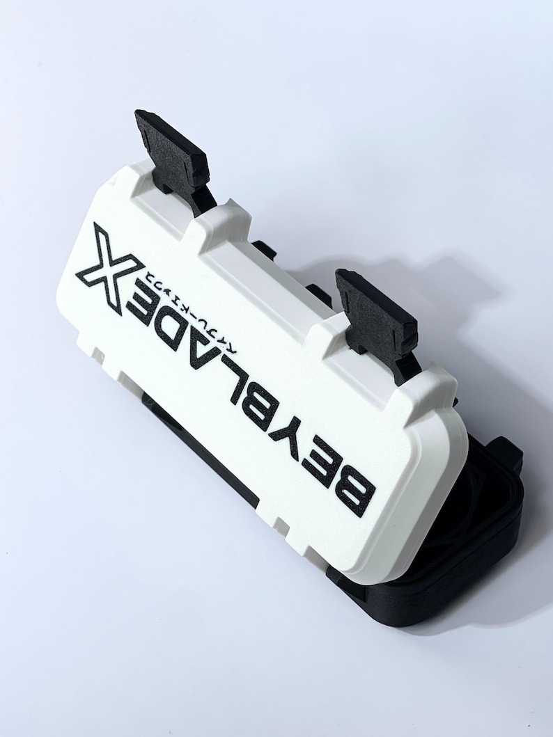 May include: A white and black plastic case with the text "BEYBLADE X" printed on the top. The case has two black handles and is partially open, revealing a black interior. The case is designed for storing Beyblade toys.