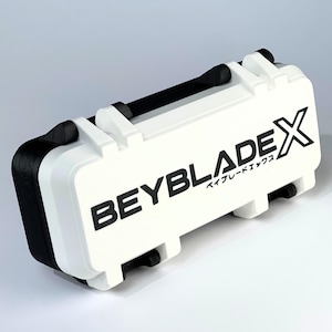 May include: White and black rectangular case with the text "BEYBLADE X" printed on the front. The case has a black handle and black accents on the sides. The case is designed to store Beyblade X products.