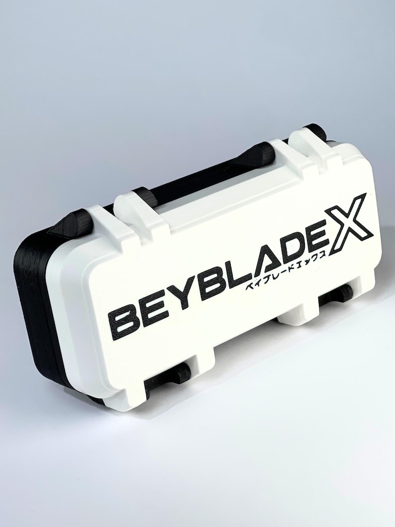 May include: White and black rectangular case with the text "BEYBLADE X" printed on the front. The case has a black handle and black accents on the sides. The case is designed to store Beyblade X products.