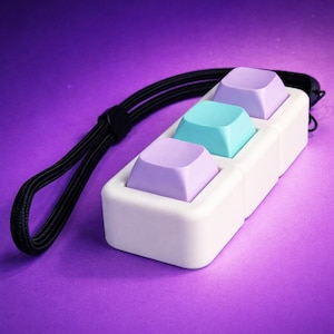 Puede incluir: Un accesorio rectangular blanco con tres teclas de teclado coloridas: dos lavanda y una turquesa. Se adjunta un cordón negro. El fondo es un degradado de púrpura.