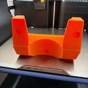 May include: A bright orange 3D printed bracket with a U-shaped cutout in the center. The object features two vertical side pillars, each containing a circular hole, and a third circular hole positioned at the base of the central arch structure.