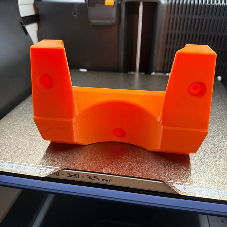 May include: A bright orange 3D printed bracket with a U-shaped cutout in the center. The object features two vertical side pillars, each containing a circular hole, and a third circular hole positioned at the base of the central arch structure.