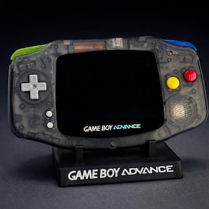 Peut inclure: Une console portable Game Boy Advance transparente sur un support noir. La console a un écran noir, un pavé directionnel et des boutons colorés. Le support et la console affichent le texte "GAME BOY ADVANCE". Le dessus de la console a des accents verts et bleus.