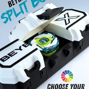 Può includere: Una scatola nera e bianca Beyblade X Split Box con un colorato giocattolo Beyblade all'interno. La scatola presenta il testo "BEYBLADE X SPLIT BOX" e "CHOOSE YOUR COLORS". Il giocattolo Beyblade è blu, verde e bianco.