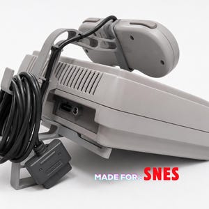 Puede incluir: Un soporte de almacenamiento de plástico gris para una consola de videojuegos y un mando. El soporte cuenta con un gancho para el cable negro enrollado del mando y una base para el mando. El texto MADE FOR: SNES está impreso en rojo abajo a la derecha.