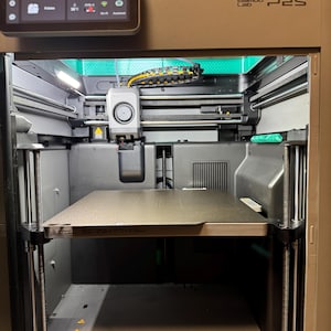 May include: A Bambu Lab P2S 3D printer with a dark gray frame and a square print bed. The top features a digital touchscreen display showing the text Un autre jour, une autre impression. The interior contains a print head, metal rods, and cables.