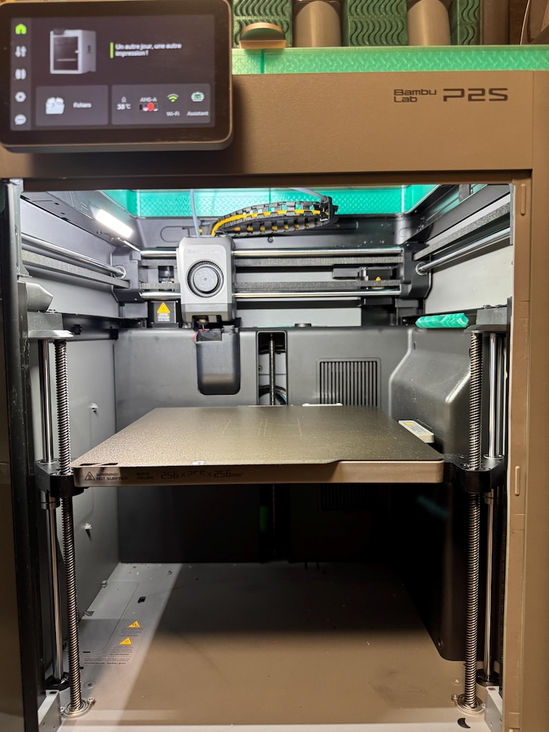 May include: A Bambu Lab P2S 3D printer with a dark gray frame and a square print bed. The top features a digital touchscreen display showing the text Un autre jour, une autre impression. The interior contains a print head, metal rods, and cables.