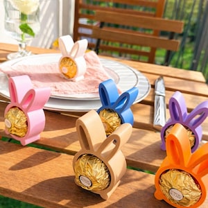 Könnte beinhalten: Ein Holztisch zeigt farbenfrohe Osterhasen-Halterungen, die jeweils ein goldverpacktes Schokoladenstück enthalten. Die Halterungen sind in verschiedenen Farben erhältlich: Rosa, Blau, Beige, Orange und Lila. Eine weiße Rose in einer Vase und ein Messer liegen ebenfalls auf dem Tisch.