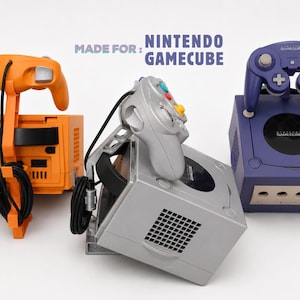 Peut inclure: Trois consoles Nintendo GameCube en orange, argent et violet, chacune associée à une manette assortie montée sur un support. Le texte MADE FOR: NINTENDO GAMECUBE est visible au-dessus des consoles. Chaque unité présente une zone de port manette.