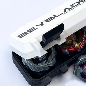May include: A white and black Beyblade storage case with the word "BEYBLADE" printed on the lid. The case is open, revealing several colorful Beyblades inside. The case is designed to hold and protect the Beyblades.
