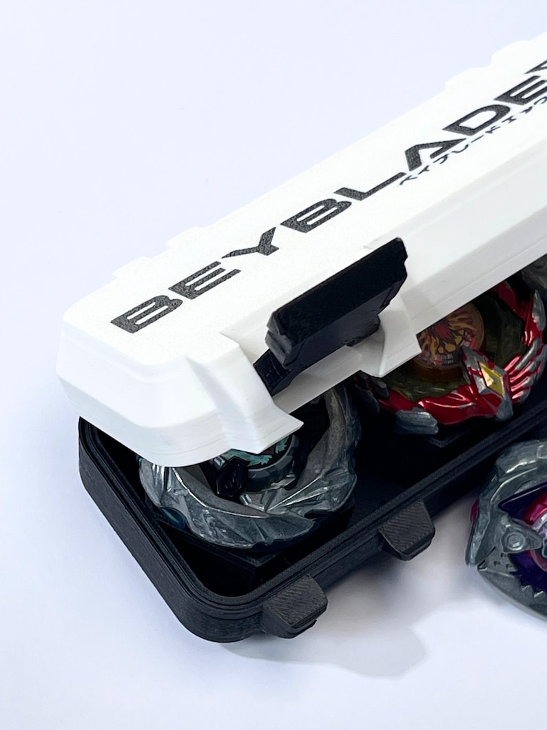 May include: A white and black Beyblade storage case with the word "BEYBLADE" printed on the lid. The case is open, revealing several colorful Beyblades inside. The case is designed to hold and protect the Beyblades.