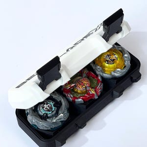 May include: A black carrying case holds three colorful spinning tops. The tops are silver, red, and gold with intricate designs. The case has a white lid with black accents and a handle.