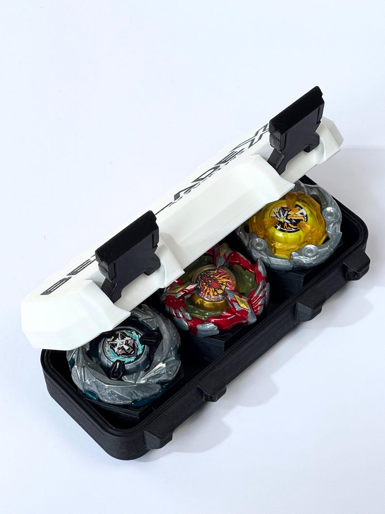 May include: A black carrying case holds three colorful spinning tops. The tops are silver, red, and gold with intricate designs. The case has a white lid with black accents and a handle.
