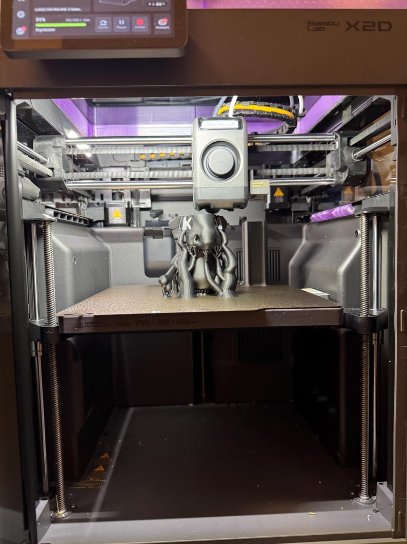 May include: A Bambu Lab X1C 3D printer with a dark gray frame and internal lighting. A black, multi-tentacled creature figure with the letter K printed on its front is positioned on the textured build plate, which is marked 256 x 256 x 256 mm.