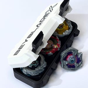 May include: A black and white case with the text "BEYBLADE X" holds several colorful spinning tops. The case is open, revealing the tops in various colors, including yellow, red, and silver. One top rests outside the case.