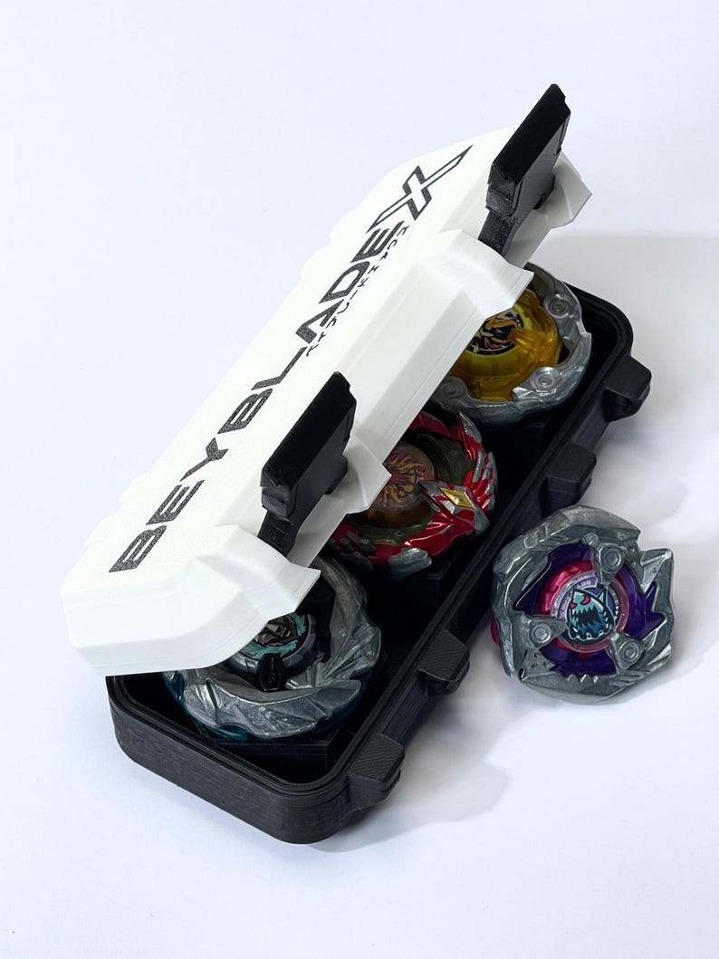 May include: A black and white case with the text "BEYBLADE X" holds several colorful spinning tops. The case is open, revealing the tops in various colors, including yellow, red, and silver. One top rests outside the case.