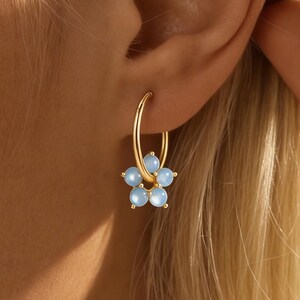 Puede incluir: Pendientes de aro dorados con un colgante de flor. La flor está hecha de cinco piedras preciosas redondas de color azul claro. Los pendientes se llevan en la oreja.