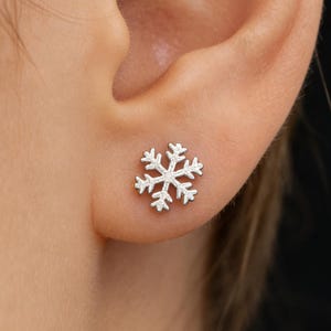 May include: A silver snowflake stud earring is shown on an ear. The earring is a detailed, six-pointed snowflake design. The earring is small and sits on the earlobe. The background is a blurred view of the person's skin and a black background.
