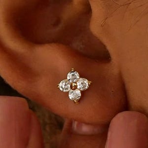 May include: A gold-colored stud earring with a four-petal flower design. Each petal features a clear, round-cut gemstone. The earring is set against the backdrop of a person's ear.