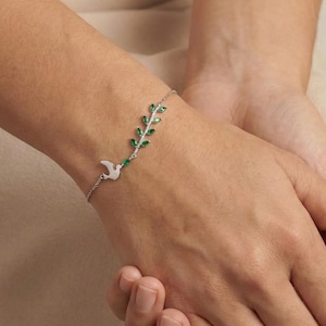 May include: A silver bracelet featuring a dove and a sprig of green gemstones. The bracelet is delicate and elegant, with a simple design. The dove is a symbol of peace, and the green gemstones add a touch of color.