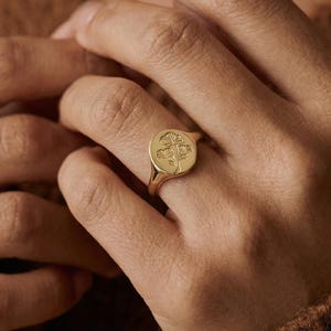 May include: A gold signet ring with a floral design. The ring features a round face with an engraved flower motif. The band is smooth and polished, and the ring is worn on a finger. The ring is a piece of jewelry.