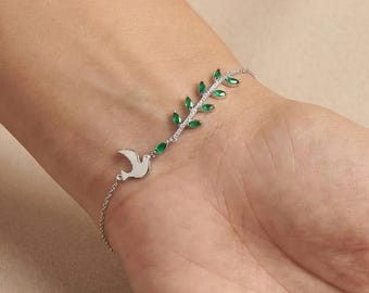 May include: A silver bracelet with a dove and leaf design, adorned with green gemstones. The bracelet is displayed on a wrist, highlighting the intricate details of the jewellery. The design incorporates a dove and a leaf with green gemstones.