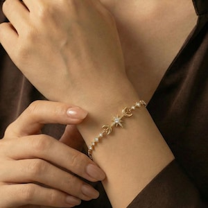 Könnte beinhalten: Goldfarbenes Armband mit einem Himmelsdesign. Das Armband hat einen Stern, Halbmonde und kleine, runde Verzierungen. Das Armband wird am Handgelenk getragen, vor einem braunen Hintergrund.