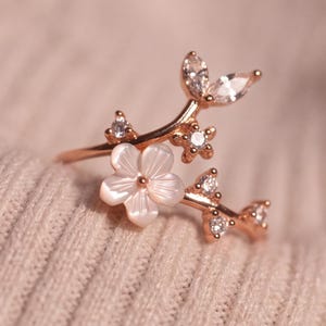 Op de afbeelding: Een roségoudkleurige ring met een takvormig ontwerp, versierd met een uitgesneden witte schelpenbloem, diverse kleine ronde doorzichtige steentjes en twee doorzichtige steentjes in marquise-slijpvorm die als blaadjes zijn geplaatst.