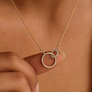 May include: A delicate gold necklace featuring two interlocking circles. One circle is plain gold, while the other is adorned with small, sparkling crystals. The necklace is displayed against a neutral background.