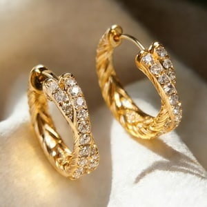 May include: A pair of gold hoop earrings with a braided design. The earrings are encrusted with small, clear gemstones. The jewelry is displayed on a white surface, with soft lighting.