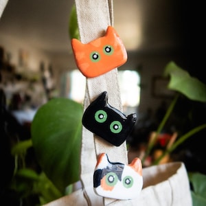 May include: Three cat-shaped magnets in orange, black, and calico designs. Each magnet has green eyes and is attached to a beige canvas bag. The magnets are decorative and add a playful touch.