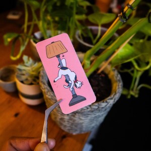 Pode incluir: Um marcador de livros rosa com um gato de desenho animado pendurado em um poste de luz. O gato é branco com manchas pretas e uma coleira vermelha. A lâmpada tem uma cúpula bege. O marcador de livros é segurado por uma pinça em frente a uma planta em vaso.