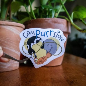 May include: A sticker with the word "Compurrrsion" in blue script. The sticker features a black and white cat, a gray cat, and an orange cat in a heart shape. The sticker is being held by tweezers in front of a potted plant.