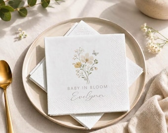 Può includere: Un tovagliolo di carta quadrato bianco con un'illustrazione stampata di un bouquet di fiori di campo nei toni del giallo, bianco e verde con una piccola farfalla. Il testo recita BABY IN BLOOM e il nome Evelynn in carattere corsivo.