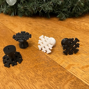 May include: Four decorative cabinet knobs with a snowflake design. Two are black, one is white, and one is black with a pedestal base. The knobs are on a wooden surface, with a green garland in the background.