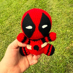 May include: A crocheted Deadpool doll in red and black. The doll features the character's distinctive mask with black eye shapes and white eye details. The doll has a cream-colored belt and is held against a backdrop of green grass.