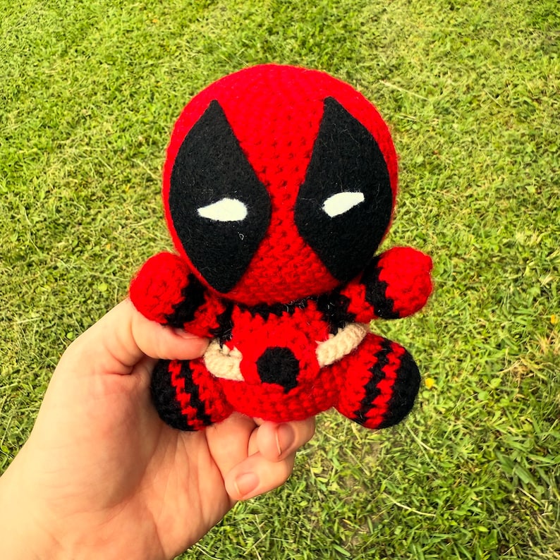 Puede incluir: Mu&ntilde;eco de Deadpool tejido a ganchillo, rojo y negro. El mu&ntilde;eco presenta la m&aacute;scara distintiva del personaje con formas de ojos negros y detalles blancos. El mu&ntilde;eco tiene un cintur&oacute;n color crema y se sostiene sobre un fondo de hierba verde.