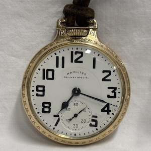 May include: A gold-toned Hamilton Railway Special pocket watch with a white face and black numerals. The watch has a small seconds dial and a brown leather strap. The watch face reads "HAMILTON RAILWAY SPECIAL".