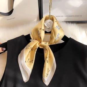 May include: A yellow silk scarf with a floral pattern and a white border. The scarf is knotted and adorned with a white flower. The scarf is hanging on a black garment.