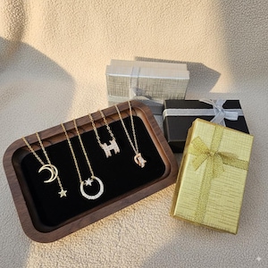 Puede incluir: Collares dorados con diseños de colgantes que incluyen una luna creciente y una estrella, un círculo con piedras incrustadas, la letra "H" y un pequeño dije. Los collares se exhiben en una bandeja de madera con cajas de regalo al fondo.