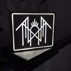 May include: A rectangular black sign with a white geometric design. The design features stylized arrows, a sunburst, and a cross. The sign is illuminated with a bright white light, creating a striking contrast against the dark background.