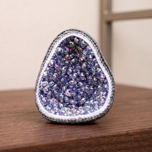 May include: A geode-shaped decorative item with a crocheted gray and white border. The interior is filled with iridescent, purple-toned crystals. The item is displayed on a dark wooden surface.