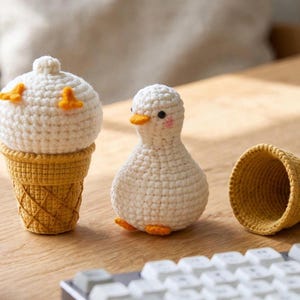 May include: A white crocheted duck figurine with an orange beak and feet, accompanied by two yellow crocheted ice cream cone shells. One cone is positioned under the duck, while the other sits empty beside it on the surface.