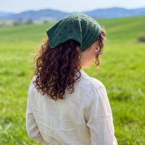 Könnte beinhalten: Ein dunkelgrünes, dreieckiges Kopftuch mit einem zarten Strickmuster und kleinen rautenförmigen Aussparungen. Der Stoff hat eine gerippte Textur an den Rändern und wird am Hinterkopf zusammengebunden getragen, wobei er den Oberkopf bedeckt.