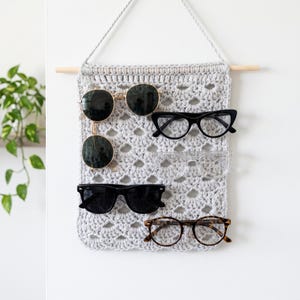 May include: A light gray crocheted wall hanging organizer featuring a wooden dowel and a hanging cord. The rectangular textile piece has an openwork pattern and holds six pairs of eyeglasses and sunglasses in various styles, colors, and frame shapes.