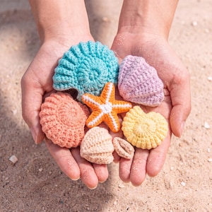 May include: A collection of six crocheted items shaped like sea life, including a bright blue spiral shell, a lavender shell, a peach-coloured shell, a yellow spiral shell, a small tan shell, and a vibrant orange and white starfish.